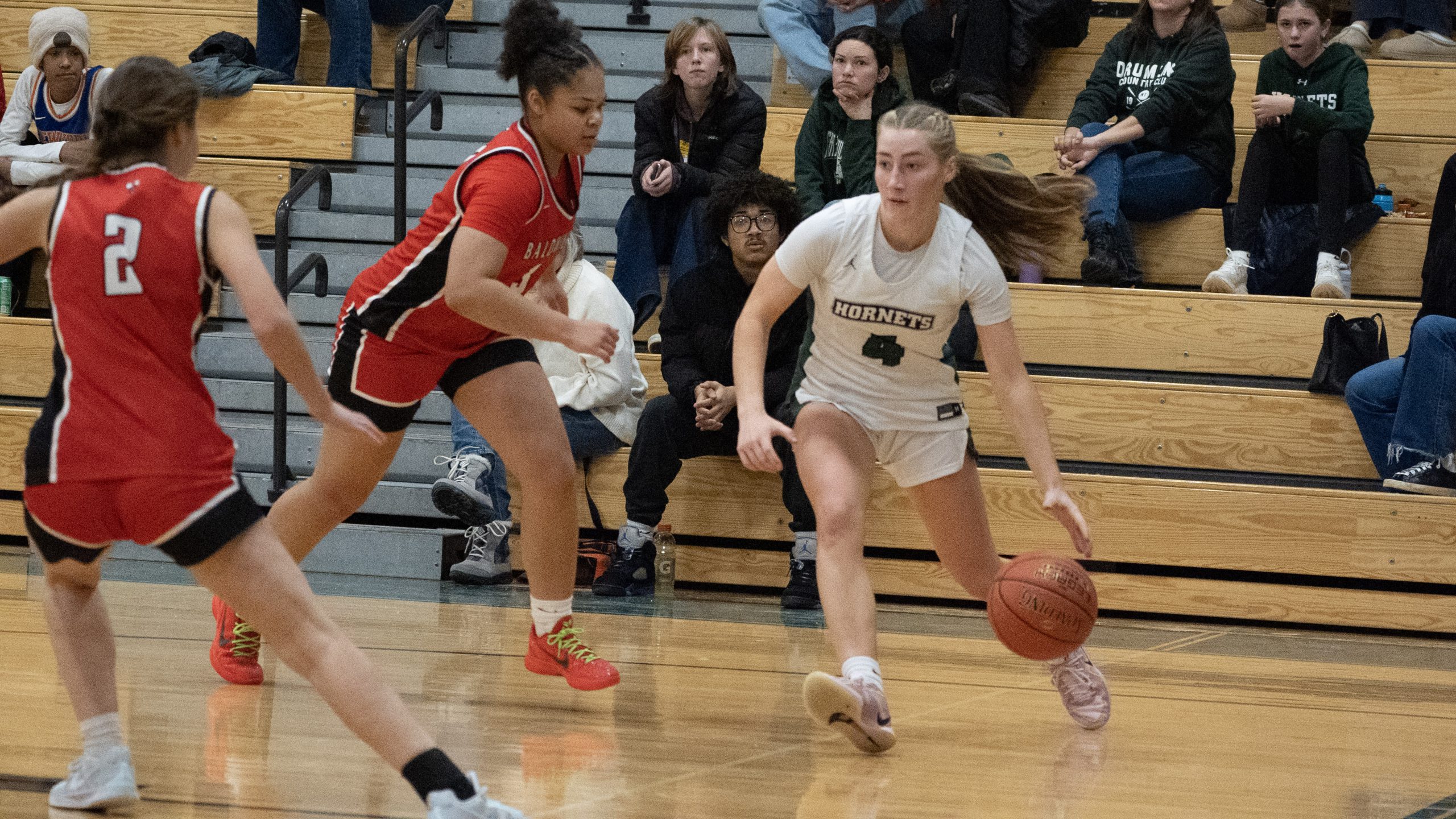One girl wearing a white basketball uniform dribbling a ball running down the court with two girls in red uniforms running toward her