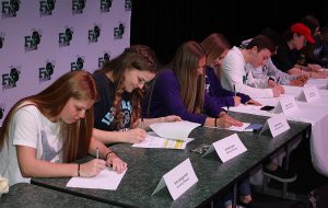 Students seated at a long table sign paperwork.