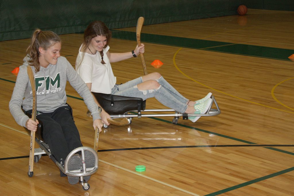 Two girls use sleds on wheels to play hockey. 