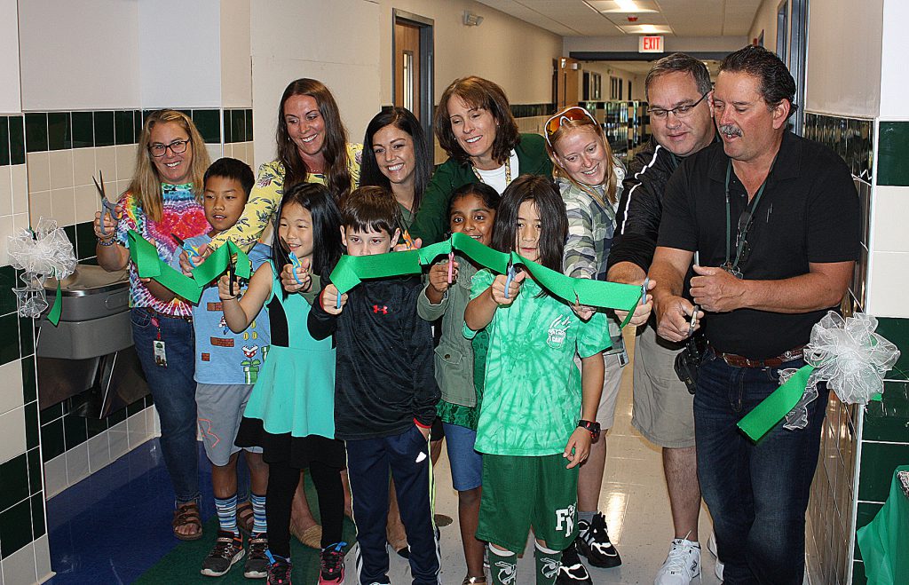 Group of people using scissors to cut a green ribbon stretched across a hallway.