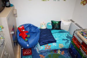 View of the room: bean bag chair with a red bear sitting in it and a small innflated bed with colorful pillows and stuffed animals on it. 