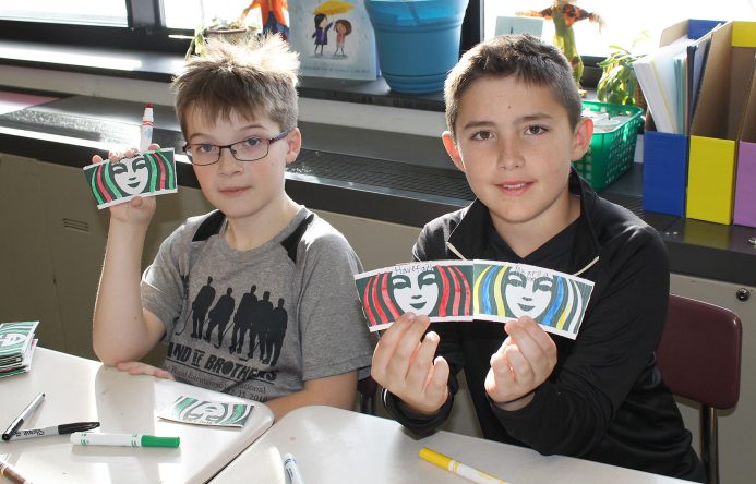 Two boys seated at desks holding up colored coffee sleeves. .