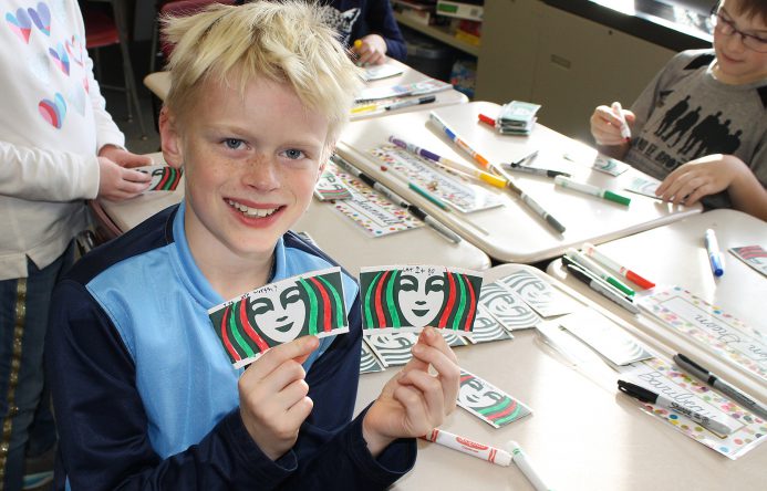 Boy seated at desk holding up colored coffee sleeves. .