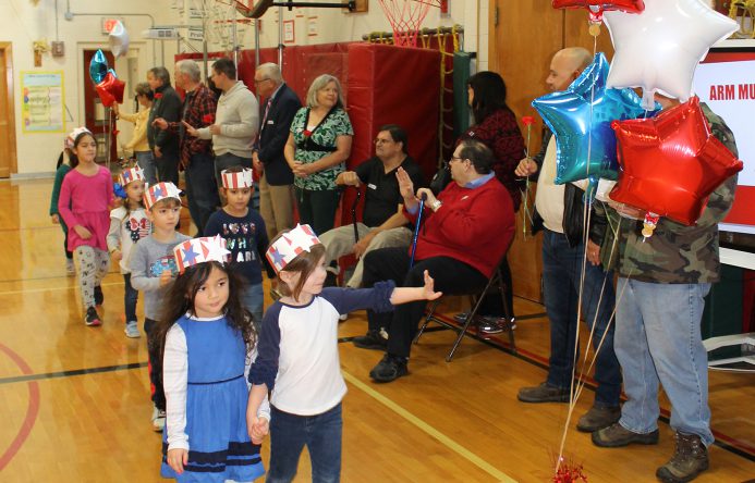 Students walking trough gym, waving at adults standing against walls. Red white and blue balloons in frame.