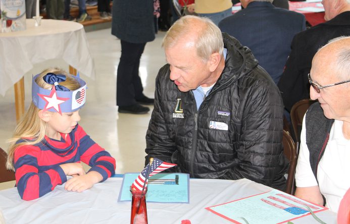 Femaile student wearing paper red, white and blue hat, eated at table talking with two men.