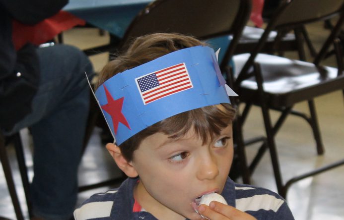 Boy waering paper red, white and blue hat eating a donut hole.