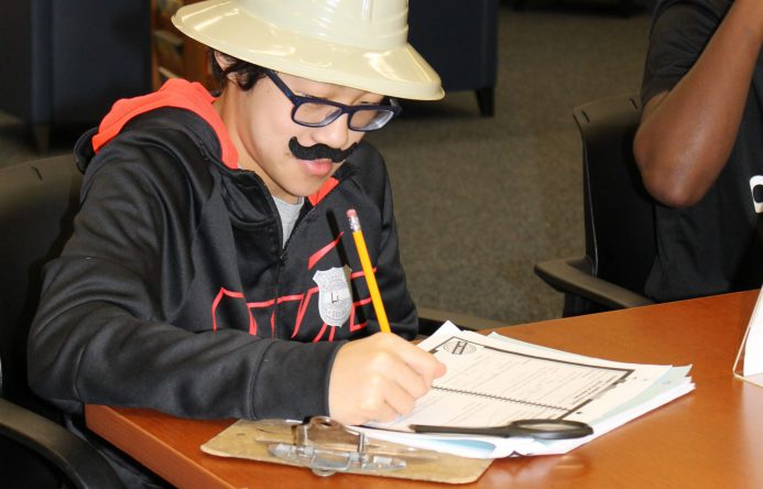 student seated at table, wearing safari hat and fake mustache, taking notes.