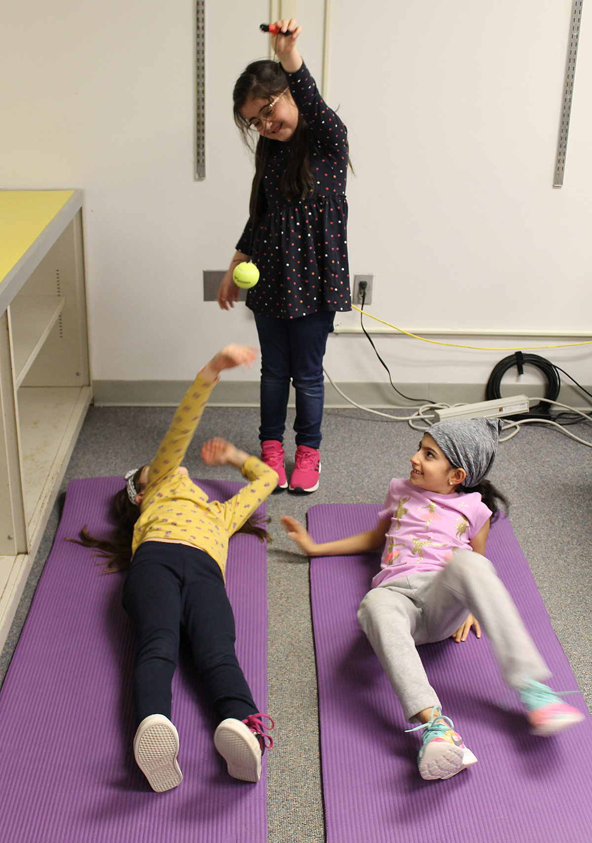 Two students laying on floor and a third student standing over them dangling a tennis ball from a string.