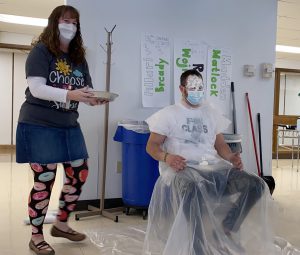 Person standing holding a pie next to a person seated with whipped cream on their face.
