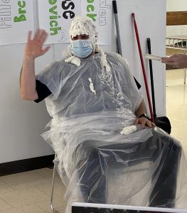 Person seated with plastic cape covering clothes and face covered in whipped cream. 