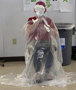 Person seated wearing a plastic cape covering their clothes. Their face is covered with whipped cream. 