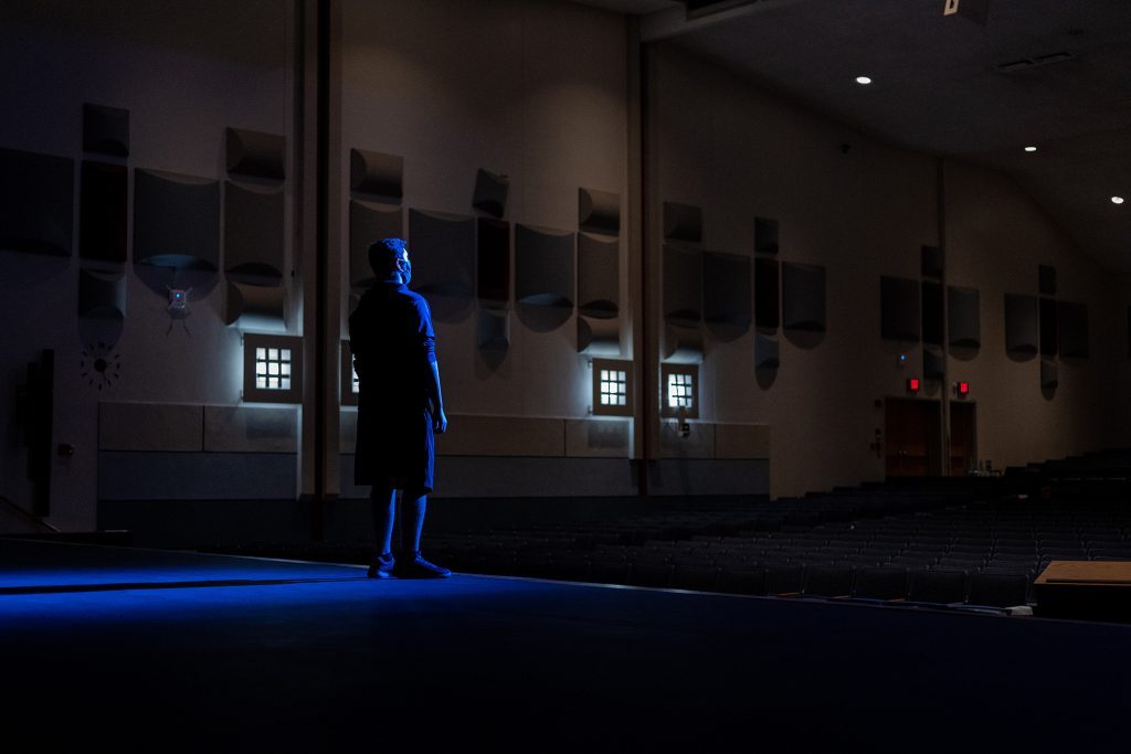 Individual wearing a face mask standing on a stage in a dark and empty auditorium. 