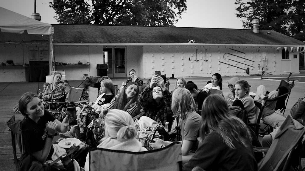 Black and white image of students seated outside in a group, taking selfies. 