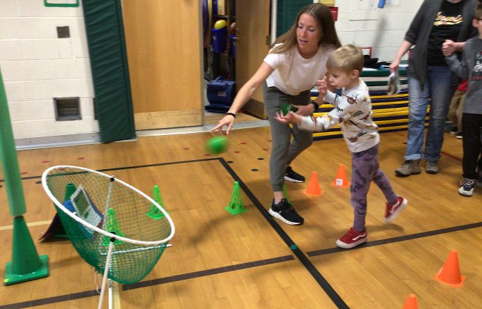 An adult helps a student throw a ball into a net.