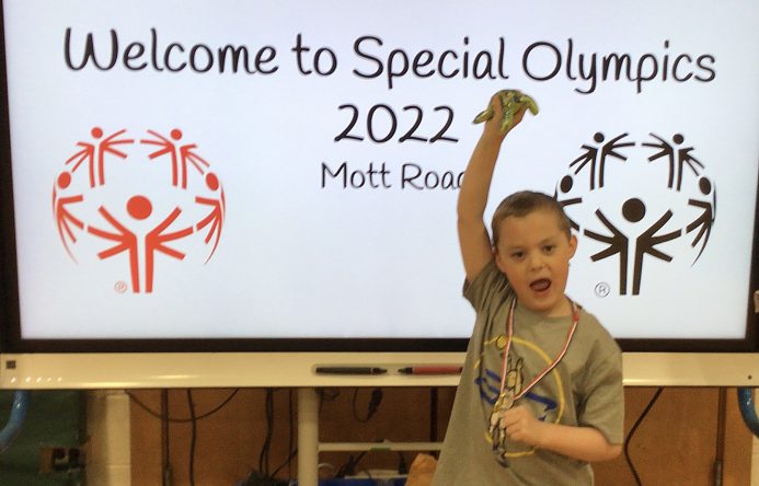 A student with his arm raised above his head stands next to a Welcome to Special Olympics 2022 sign.