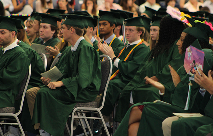 Graduates sitting in chairs and clapping at graduation.