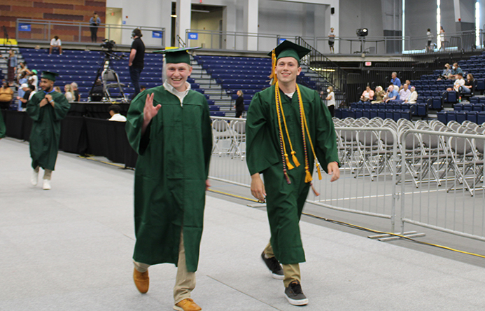 Two students smiling and waving as they walk to the graduation stage.