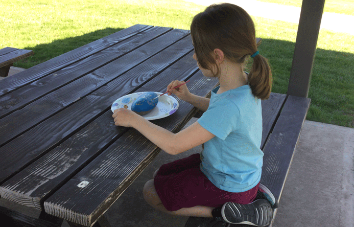 Student sitting at a picnic table and painting a rock.