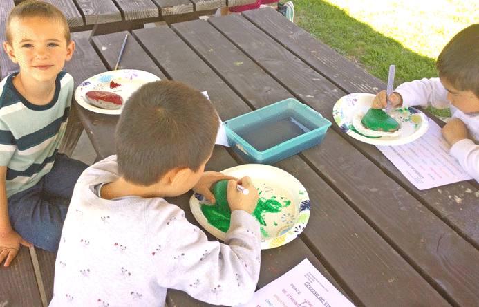Three students painting rocks at a picnic table.