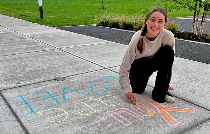 A female student is on her knee writing you are loved on the sidewalk