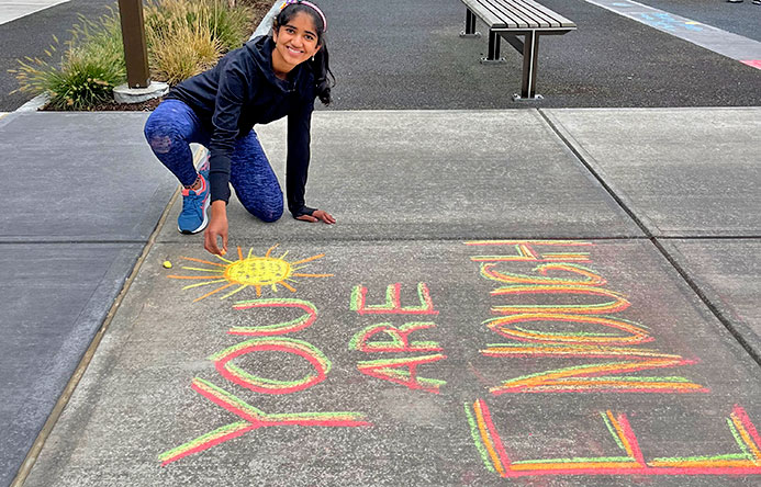 A young female student chalks a message onto the concrete outside of their school