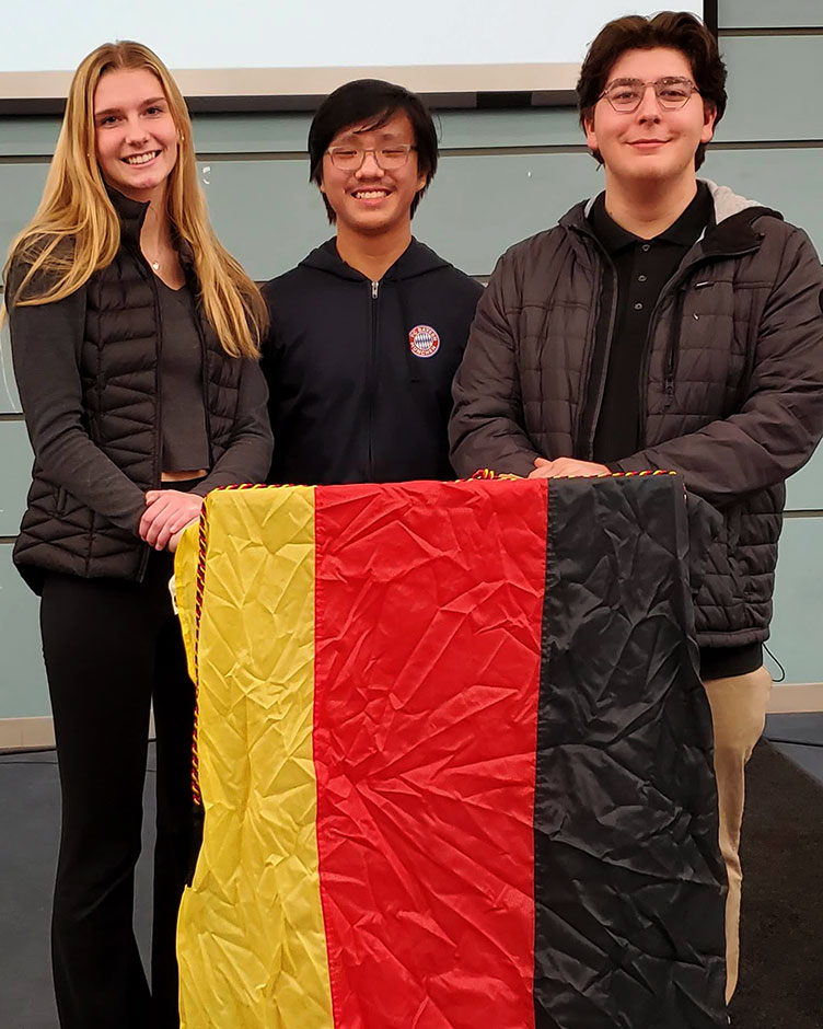 Three high school students who study German stand together for a photo. They are holding the German flag.