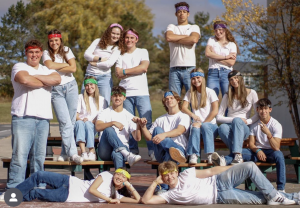 14 high school students dressed in white shirts and colored bandanas pose on picnic tables outside.