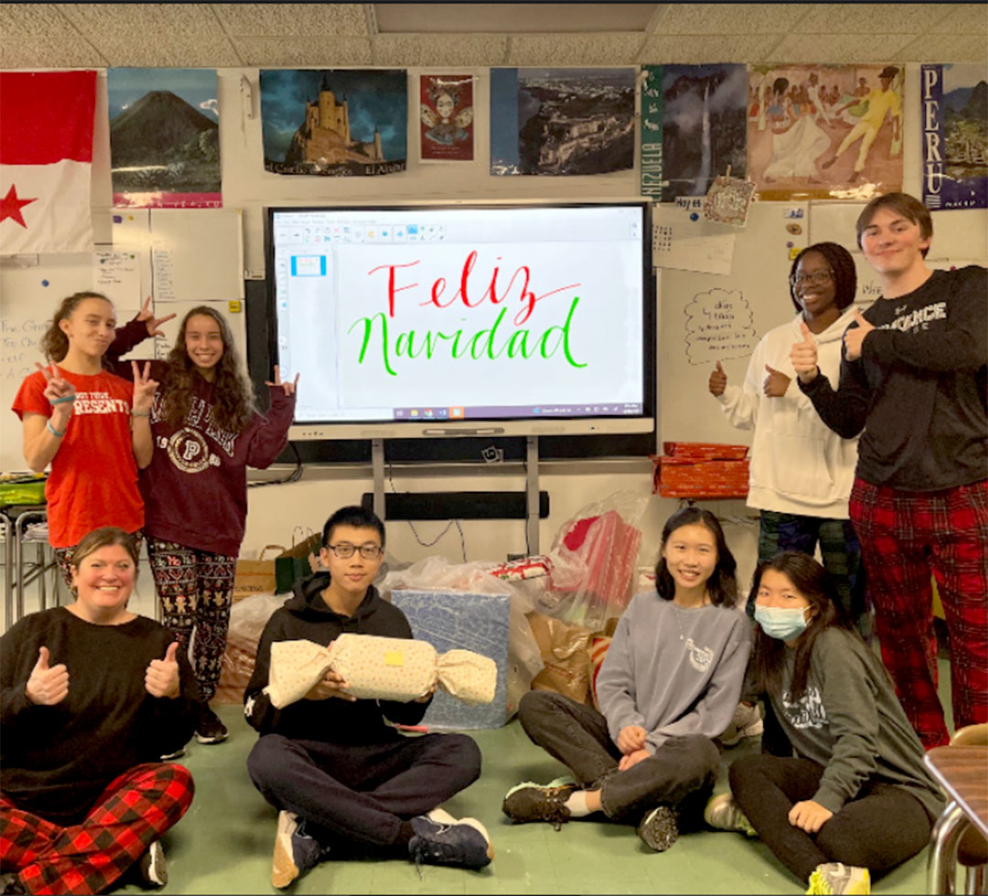 A group of students sit and stand together in a classroom for a photo. Some of them are holding wrapped gifts