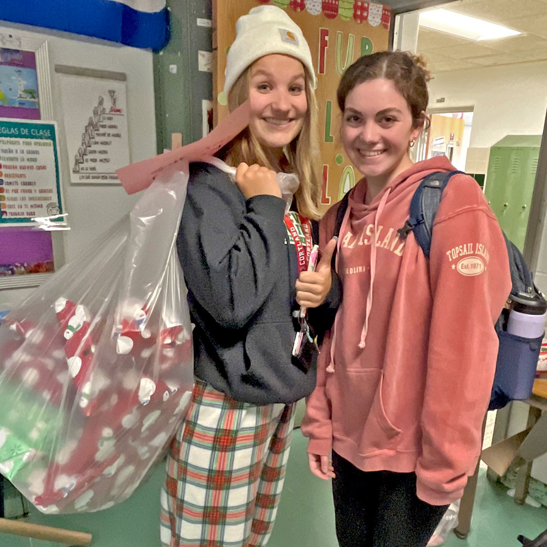 Two high school students stand inside a classroom. One of them is holding a bag of gifts.