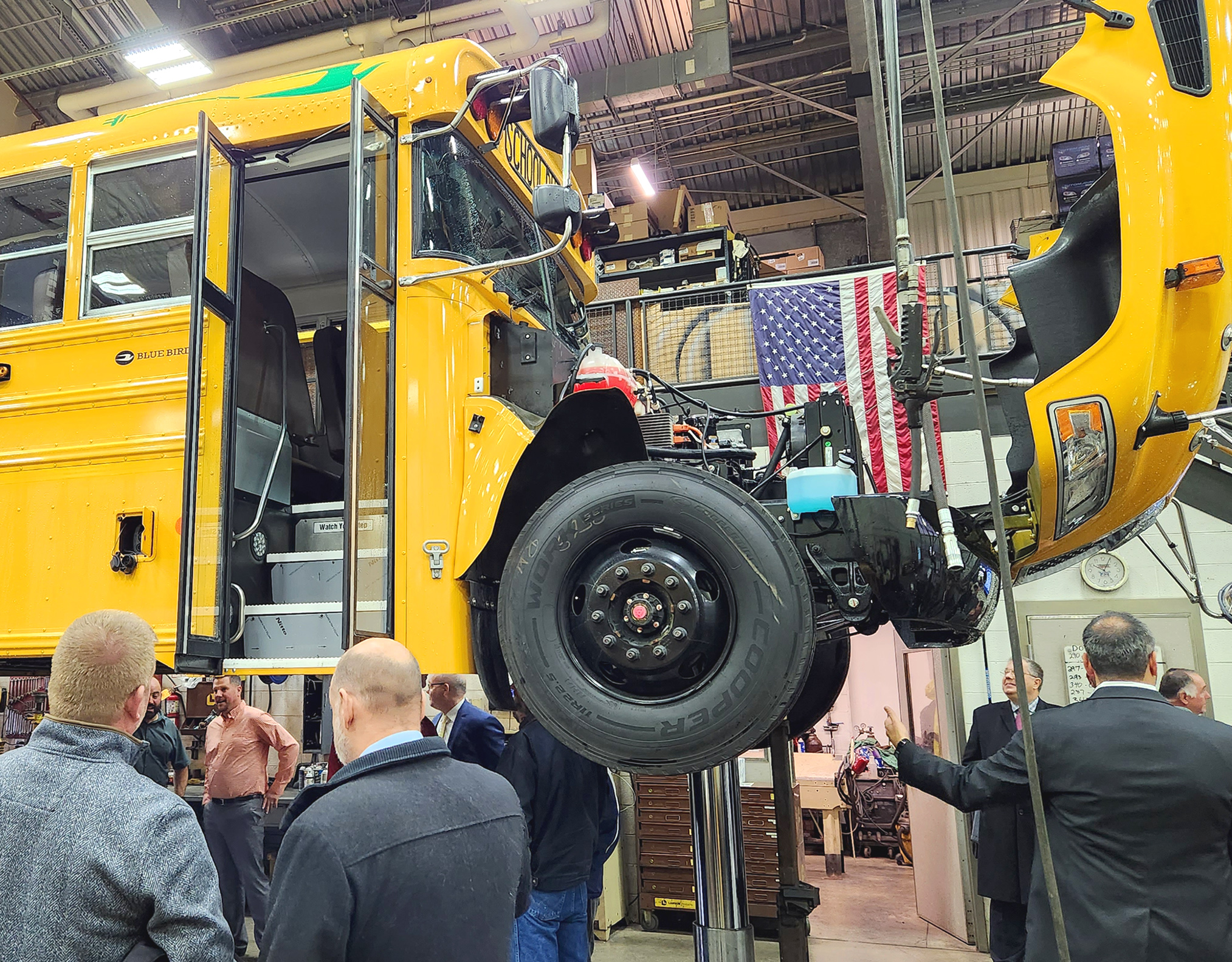 Several people are viewing an electric bus that is raised up by a lift.