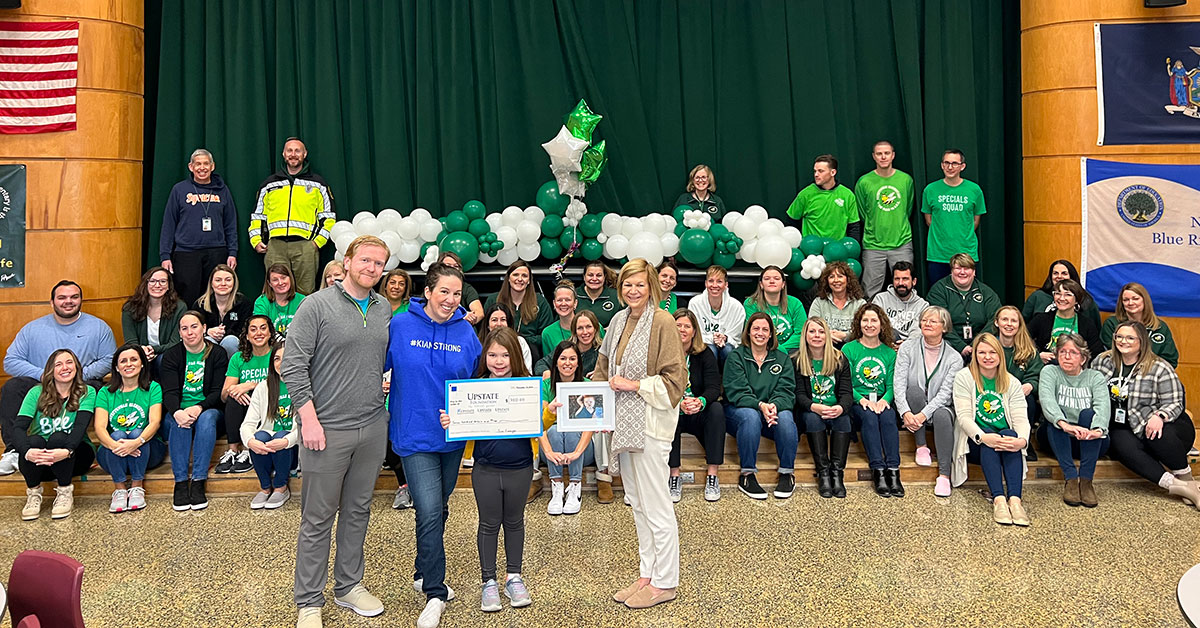 Dozens of school staff members are sitting and standing on the school's stage and June, her parents, and Upstate University Hospital Professor of Pediatrics Irene Cherrick, MD are standing on the floor.