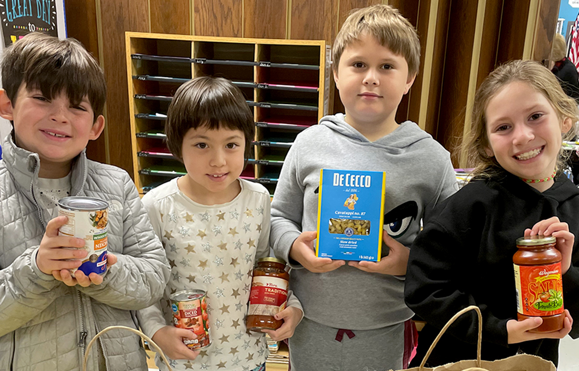 Four elementary students stand together. They are each holding a non-perishable food item.