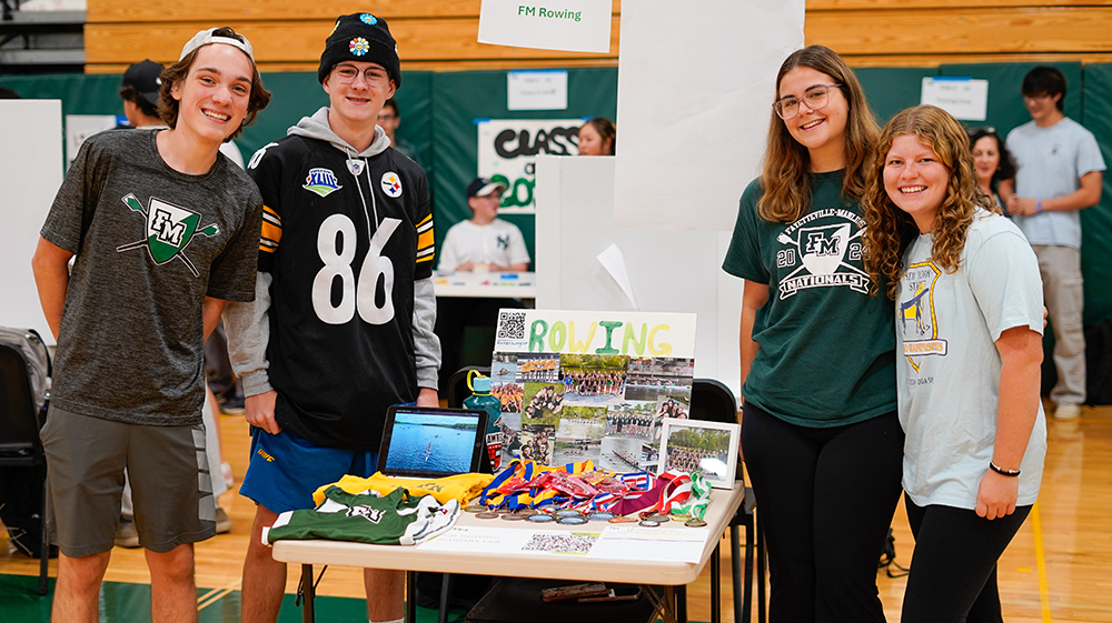 Four high school rowing students surrounding a display table about the rowing program.