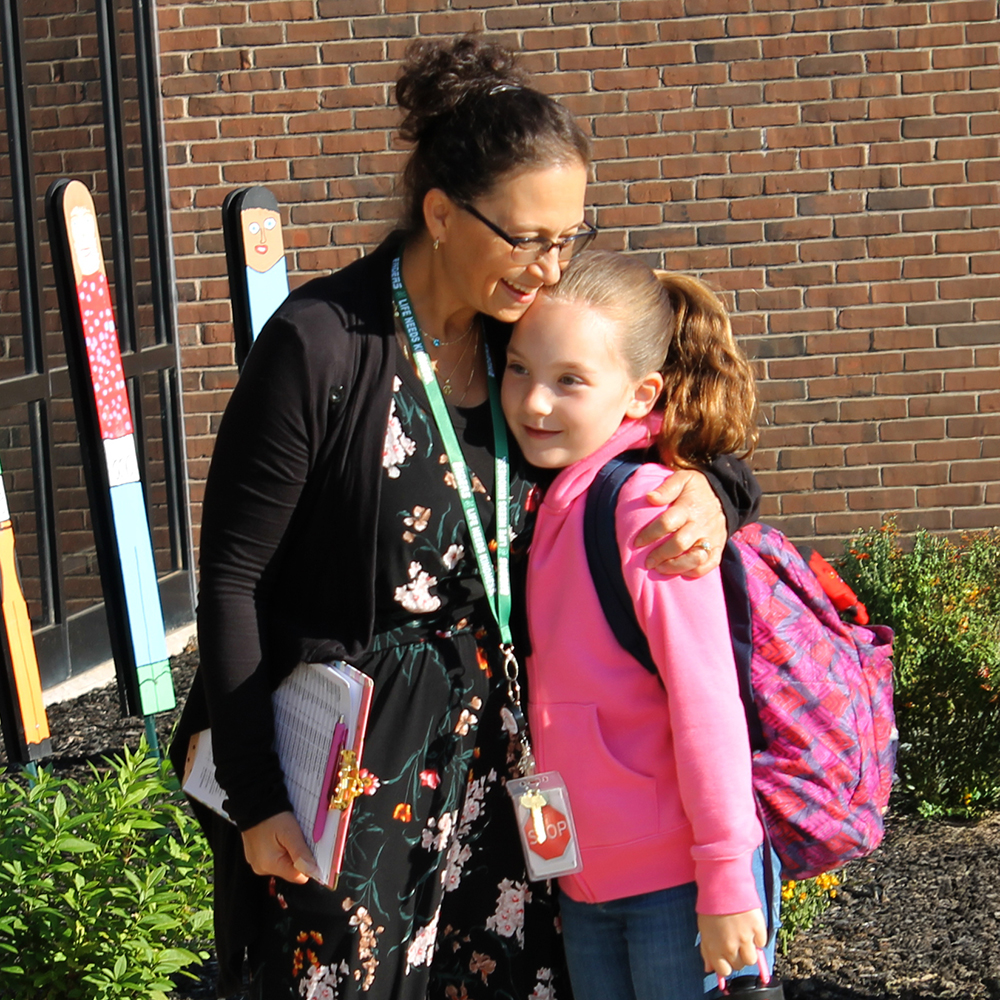A teacher hugs an elementary student outside the entrance to school.