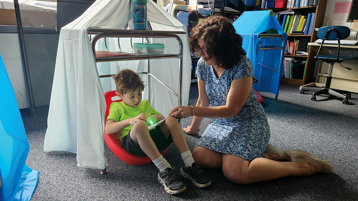 A teacher sits on the floor assiting a student reading in a fort.
