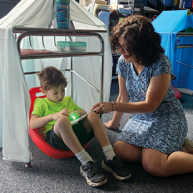 A teacher sits on the floor reading with a student in a desk fort with curtains.