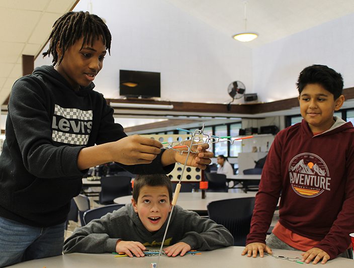 One middle school student balances wire strands while two others watch.