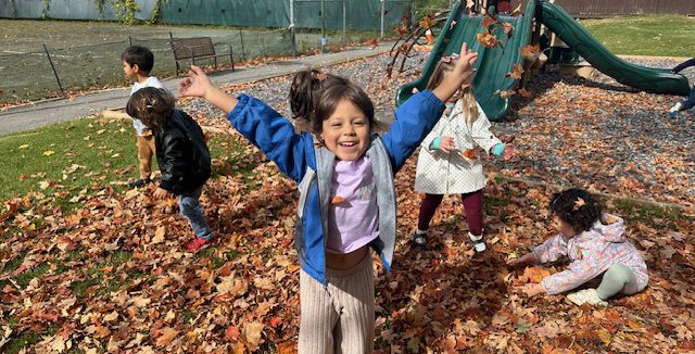 A pre-k student joyfully throwing autumn leaves in the air on the playground.