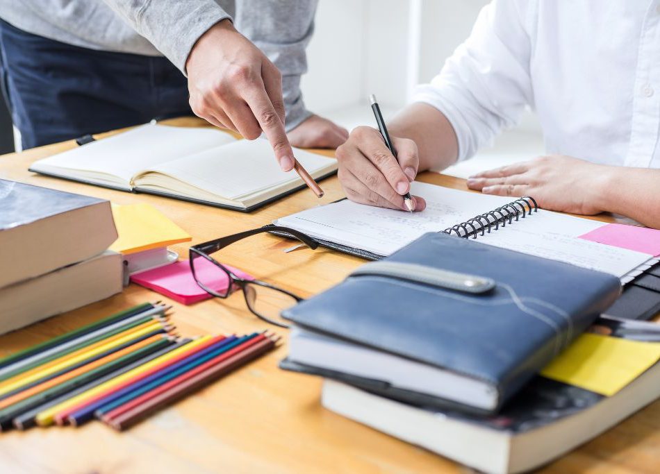 The hands of an adult indicating instructions to the hands of a student writing in a spiral notebook.