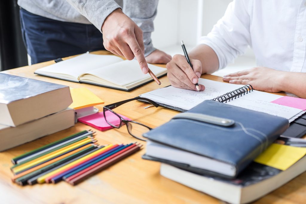 The hands of an adult indicating instructions to the hands of a student writing in a spiral notebook.