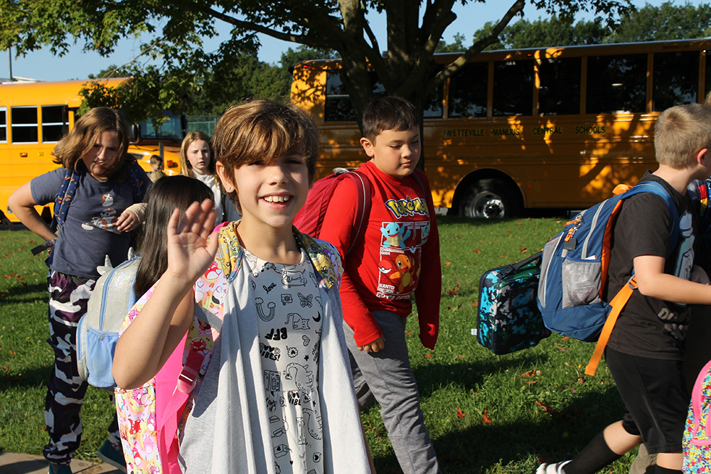A student with busses behind her waves as she has just arrived to school.