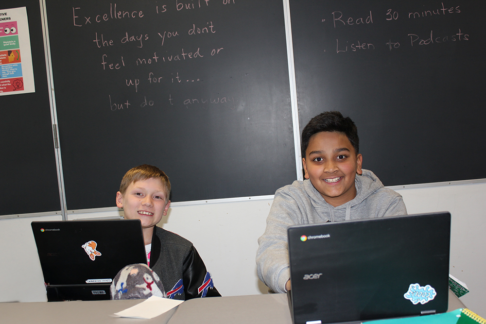 Two middle school students smile from behind laptops.