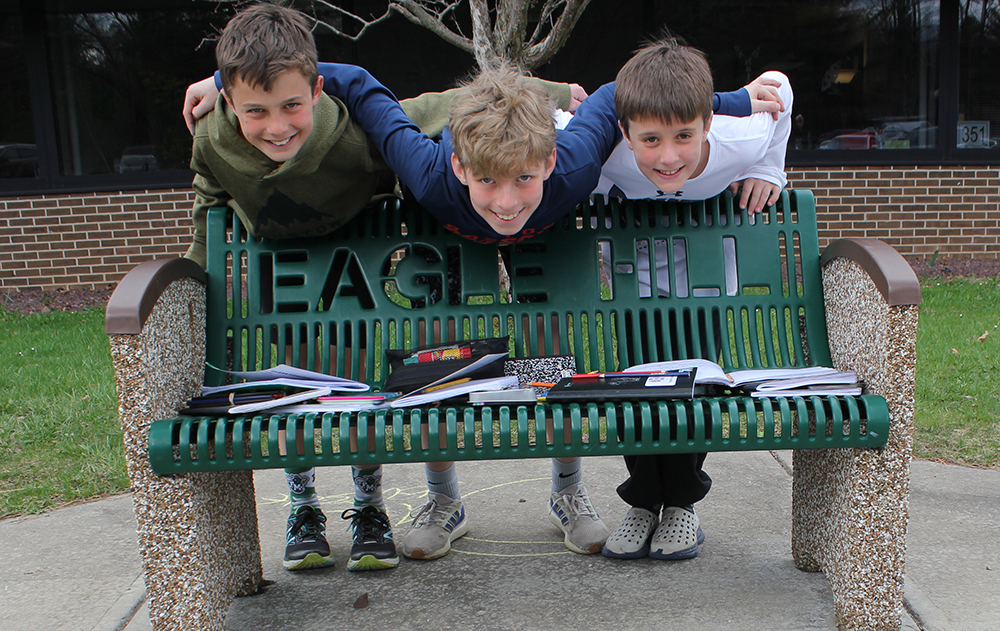 Three middle school boys peer over a bench that says "Eagle Hill".
