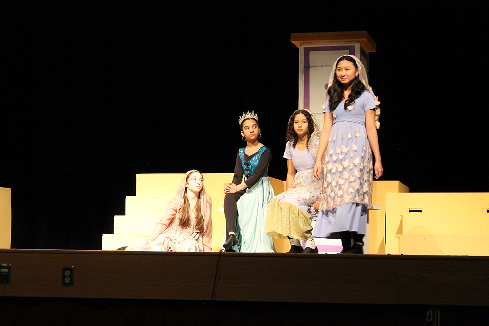 Four students in princess costume on stage acting a scene from a play.