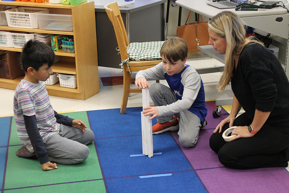 A teacher overlooks a student building a tower with another student looks on.