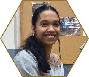 A high school student smiles in front of a corkboard in the woodshop classroom.