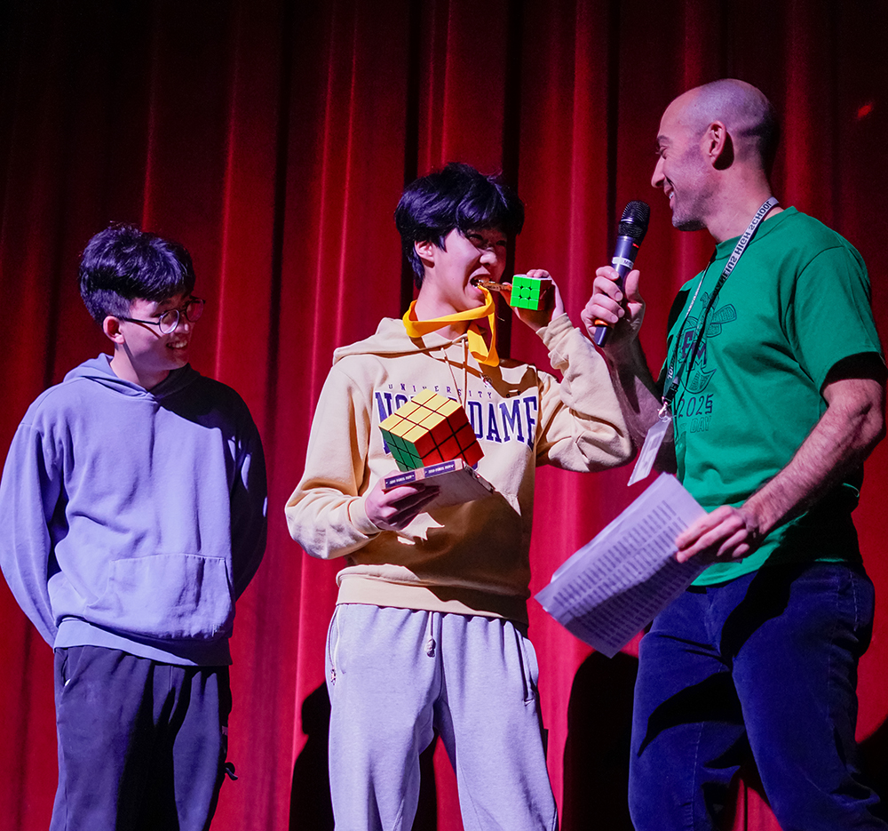 A high school student bites his medal in front of a judge on stage while another student looks on.