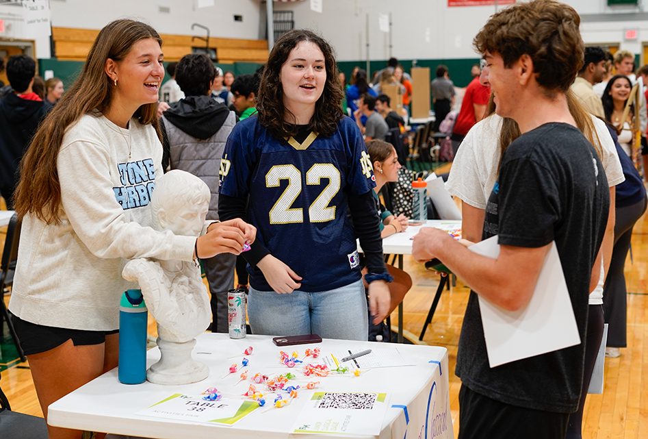 Two high school students speak with another student at their display table in a crowded gym.