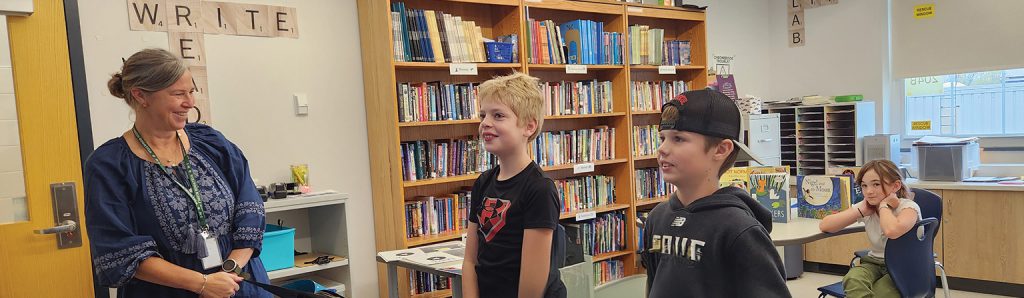 Two middle schoolers present to a smiling teacher while another student looks on from her seat.