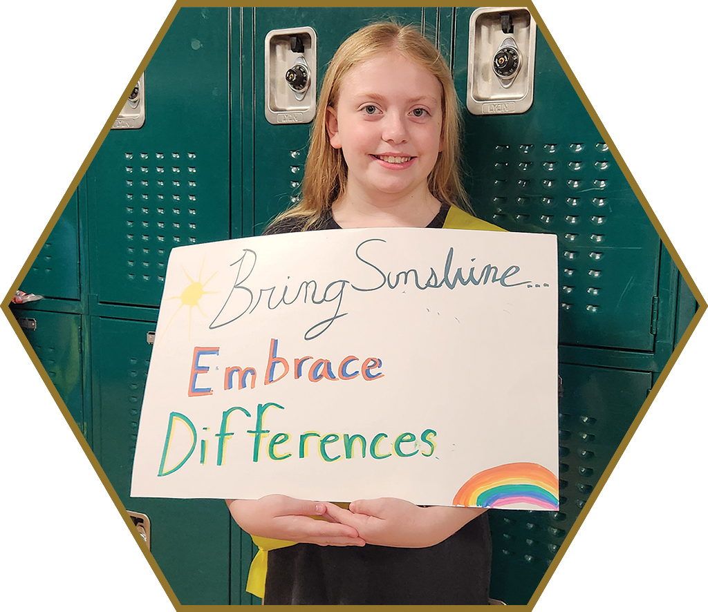 A middle school student poses with a poster she created that says "Bring sunshine... embrace differences."